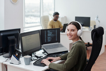 Poster - Happy young female it-engineer in headphones sitting by desk in front of computer monitor and laptop and looking at camera in office