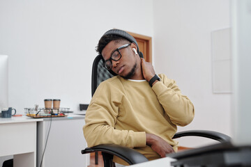Poster - Young African American man in casualwear, eyeglasses and earphones doing relaxing exercise for neck by his workplace in office