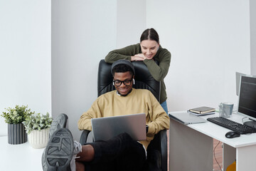 Poster - Two happy young multiracial software developers looking at screen of laptop on knees of African Americaan man sitting in armchair
