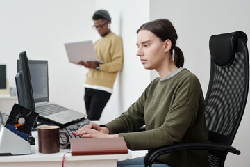 Poster - Young serious female programmer in casualwear looking at computer screen while decoding data by workplace against coworker