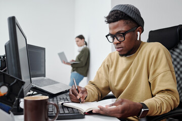 Poster - Young serious black man in casualwear making notes by workplace while sitting against female coworker with laptop standing by wall
