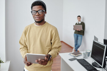 Poster - Contemporary young male programmer with tablet looking at camera against female coworker with laptop networking by wall
