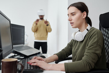 Poster - Young serious female programmer in casualwear decoding data and looking at computer screen against male coworker in vr headset