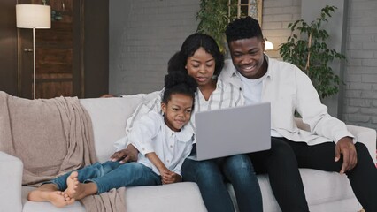 Happy African family and cute kid child girl daughter using laptop looking at computer screen enjoying watching funny social media video having online video call conference relaxing on sofa at home