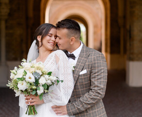 Front view of elegant bridegroom in suit, which standing and embracing bride. Happy  woman  in white wedding dress, holding beautiful flower bouquet, touching groom face and posing 