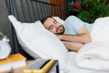 Side View Young Peaceful Man In Casual Blue T-shirt Lying In Bed Hands Folded Under Head Close Eyes Rest Relax Spend Time In Bedroom Lounge Home In Own Room House Wake Up Dream Real Estate Concept