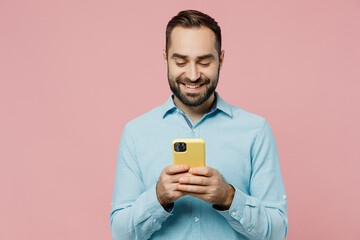 Wall Mural - Young smiling happy caucasian man 20s in classic blue shirt hold in hand use mobile cell phone chatting online isolated on plain pastel light pink background studio portrait. People lifestyle concept.