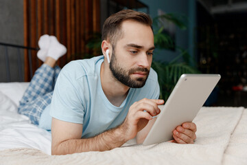 Wall Mural - Full body confident young man in blue t-shirt lying on bed listen music in earphones use tablet pc computer rest relax spend time in bedroom lounge home in own room house dream. Morning mood concept.