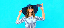 Portrait Of Beautiful Happy Smiling Woman Taking Selfie By Smartphone Wearing Black Round Summer Hat, White Striped Shirt On Blue Background