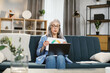 © Sofiia Shunkina - Serious adult grey-haired woman in headset having video conference on laptop while working from home. Attractive pleasant mature female sitting on sofa and showing graphs and charts.
