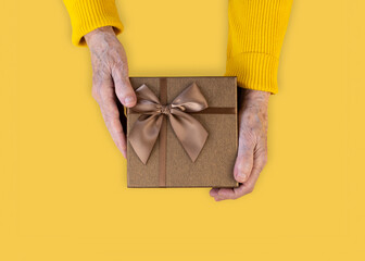 Hands of elderly woman in yellow sweater hold a gift box tied with a ribbon. Day of Elderly People concept.