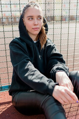 Stunning girl in black pullover with hood looking aside hand while resting outdoor. Inspired long-haired female model with big green eyes k sitting on the ground