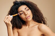 © SHOTPRIME STUDIO - MAKEUP ARTIST CONCEPT. Enjoyed tanned awesome curly Latin lady hold makeup brush after makeup visage ready to party posing isolated on pastel beige background look aside. Closeup cosmetic offer