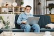 © sofiko14 - Handsome mature gentleman using laptop for video conversation sitting on sofa in living room at home. Senior bearded man freelancer in casual wear sitting on couch and having online meeting.