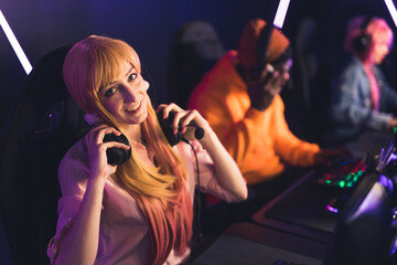 Adorable caucasian long-haired blonde gamer girl looking at camera, smiling, and putting her professional headset on. PC bang environment. High quality photo