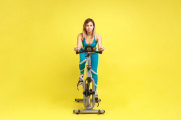 Attractive confident young woman riding on spinning bike, cycling on gym machine bicycle, motivation and sporty goal concept, wearing blue sportswear. Indoor studio shot isolated on yellow background.