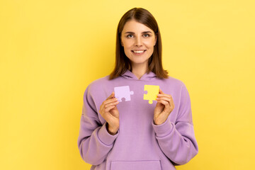 Smiling woman holding and showing two jigsaw puzzle pieces, matching and connecting puzzles, solving problems with good result, wearing purple hoodie. Indoor studio shot isolated on yellow background.