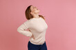 Portrait of blond woman screaming in acute pain and holding sore back, risk of kidney stones disease, pinched nerve, wearing white sweater. Indoor studio shot isolated on pink background.