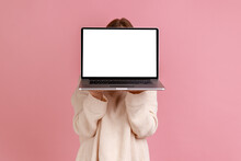 Portrait Of Blond Woman Holding Laptop In Hands, Hiding Her Face Behind Notebook With Empty Display With Copy Space, Wearing White Sweater. Indoor Studio Shot Isolated On Pink Background.