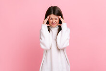 Woman Frowning And Clasping Sore Head, Suffering Intense Headache, Having Migraine, Fever Symptoms, Wearing White Casual Style Sweater. Indoor Studio Shot Isolated On Pink Background.