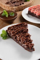 Wall Mural - Piece of Chocolate Brownie Slice with Strawberry and Chocolate Icing. Wooden table with mint and chocolate chips in the background