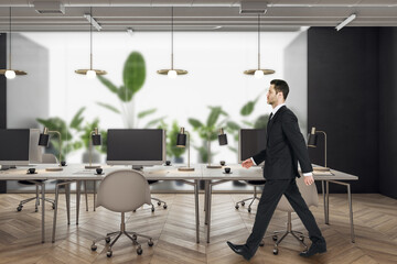 Poster - Thoughtful young man walking in modern coworking office interior with equipment, furniture, decorative plants and matte glass partition. Workplace concept.