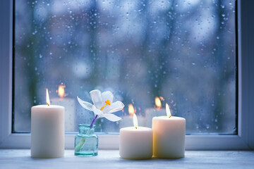 Wall Mural - Burning candles, white flower in bottle on windowsill, abstract blurred raindrops glass background. spring season. Melancholy cozy mood. Relax, harmony, meditation, life balance concept. rainy weather