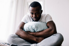 Emotionally Stressed Man Sitting Cross-legged With Pillow In Bedroom At Home