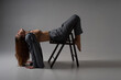 A model girl poses professionally for a photographer on a chair in complex acrobatic recumbent poses. In a formal suit and a sexy unbuttoned gray jacket, barefoot