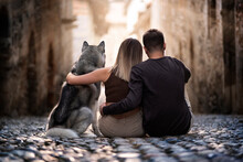 Back View Of Two Young People And Their Beloved Wolf Dog Sitting And Hugging In An Old Town Street Staring At The Sun, Piemonte