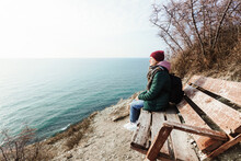 A Young Woman Traveler With A Backpack Sits On A Bench On The Edge Of A Cliff Overlooking The Sea And Sky.