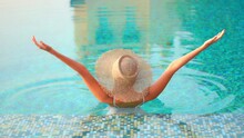 Woman Wearing Hat Standing In Warm Clear Swimming Pool With Colorful Blue Tile. Asian Lady Relaxing In Resort Pool Stretches Arms Over Head And Takes In Sunshine Of Tropical Island Paradise.
