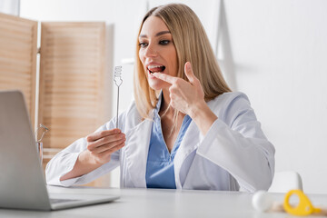 Speech therapist holding tool and pointing at mouth during video call on laptop.