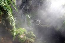 Sunlight Piercing Through Mist In A Tropical Rock Garden With A Waterfall And Pond And Mossy Green Plants Being Sprayed. No People, Southeast Asia.