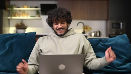 Wall Mural - Cheerful Indian man wearing casual hoodie sitting on the sofa and using laptop at home. Inspired male entrepreneur typing on keyboard and looks aside with pleasure smile, taking a break and resting