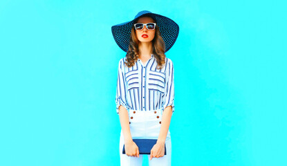 Portrait of beautiful woman model wearing black round summer hat, handbag and white striped shirt on blue background