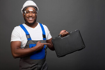 Strong black male auto mechanic by profession advertises professional tools in convenient box. Cheerful African-American man in robe and protective helmet demonstrates box with tools for auto mechanic