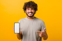 Positive Happy Indian Man Showing Smartphone With White Empty Display, Points Finger On It, Bragging With Application. Indoor Studio Shot Isolated On Yellow Background