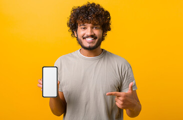 Positive happy Indian man showing smartphone with white empty display, points finger on it, bragging with application. Indoor studio shot isolated on yellow background