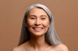Happy beautiful middle-aged grey haired Asian woman posing in studio on brown background. Cheerful mature korean lady with fresh and hydrated skin looks at the camera and laughing