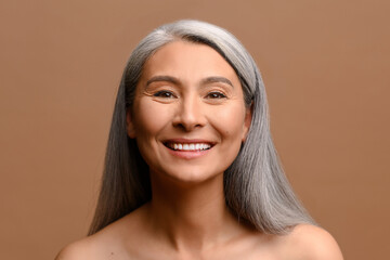 Poster - Happy beautiful middle-aged grey haired Asian woman posing in studio on brown background. Cheerful mature korean lady with fresh and hydrated skin looks at the camera and laughing