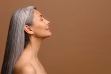 Side View At Sensual Naked Middle-aged Beautiful Asian Woman With Long Grey Hair, Her Eyes Closed, Body And Face Care Concept. Charming Mature Lady Stands In Profile Isolated On Brown Background