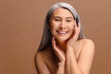Enchanting Topless Middle Aged Asian Woman Looks At The Camera And Laughing Happily, Isolated On Brown Background, Charming Korean Lady With Grey Hair Holding Face Gently. Skin Care And Self Love