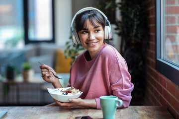 Beautiful young woman listening music with headphones while having healthy breakfast in the living room at home.