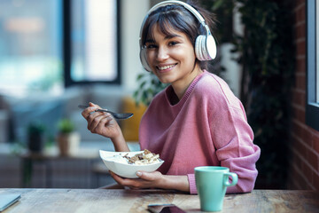 Beautiful young woman listening music with headphones while having healthy breakfast in the living room at home.