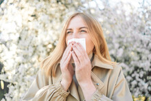 Caucasian Woman Feels Allergy, Holds White Tissuue, Stands Near Tree With Blossom. Allergy Concept