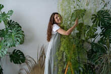 Smiling Young Woman Gardener In Gray Sundress Stands On Stepladder Hugging Lush Houseplant In Her Flower Shop. Greenery Office, Indoor Cozy Garden, Modern Green Space For Relax. Love Of Plants. 