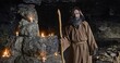 © procinemastock - Medieval monk, with a staff in a hooded place, stands in a dark cave with glowing candles in the background.
