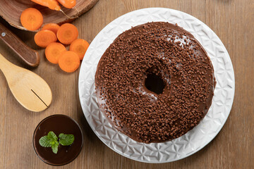 Wall Mural - Brazilian carrot cake with chocolate frosting on wooden table with carrots in the background. Top view.