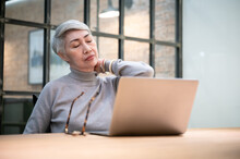Office Syndrome Concept. Mature Asian Business Woman Wears Glasses Using Laptop Computer Sit At Workplace Desk. She Stretch Her Arm And Relax Neck.